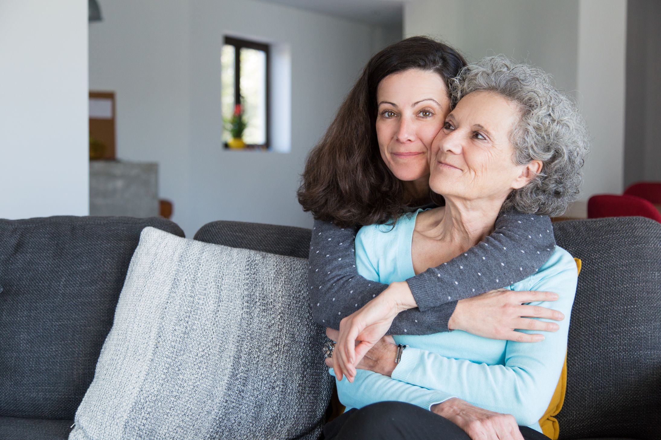 Adult daughter embraces elderly mother, who is seated on a sofa, from behind.